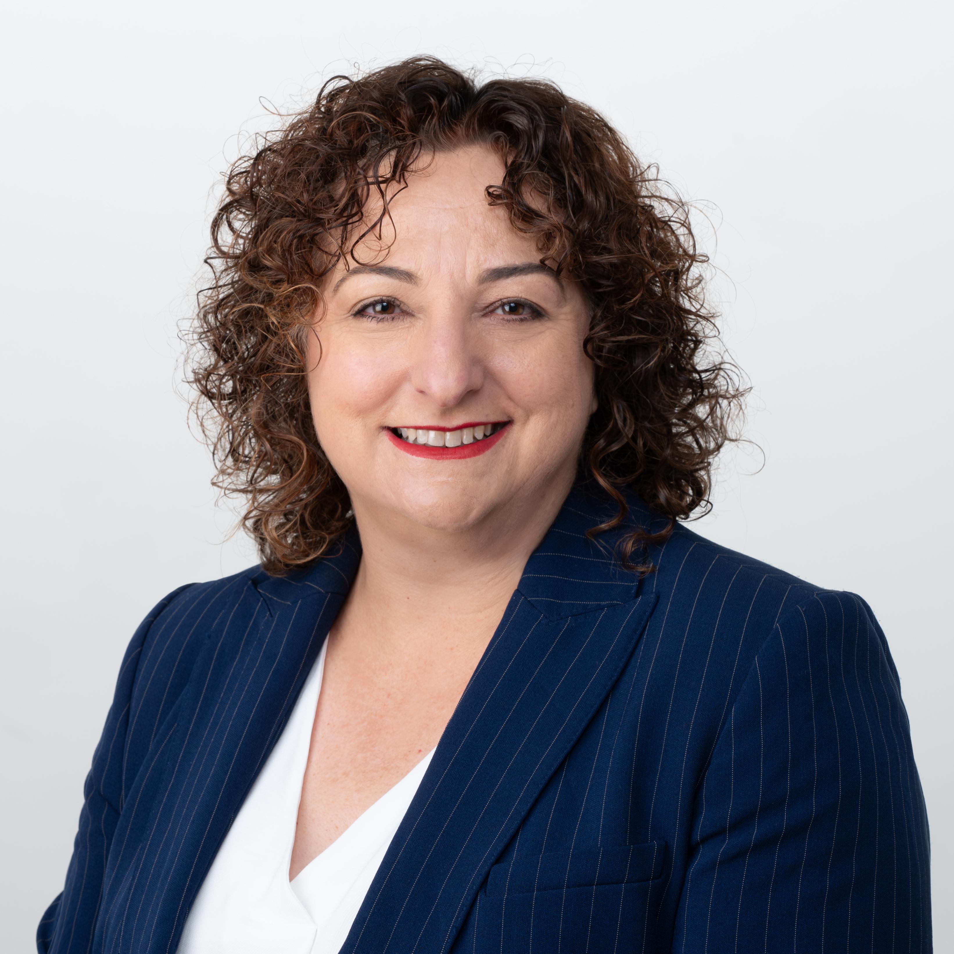 Photograph of Ms Anna Storti, Board Member of the Veterinary Practitioners Registration Board of Victoria. Anna has curly brown shoulder-length hair. She is wearing a white shirt and blue blazer.